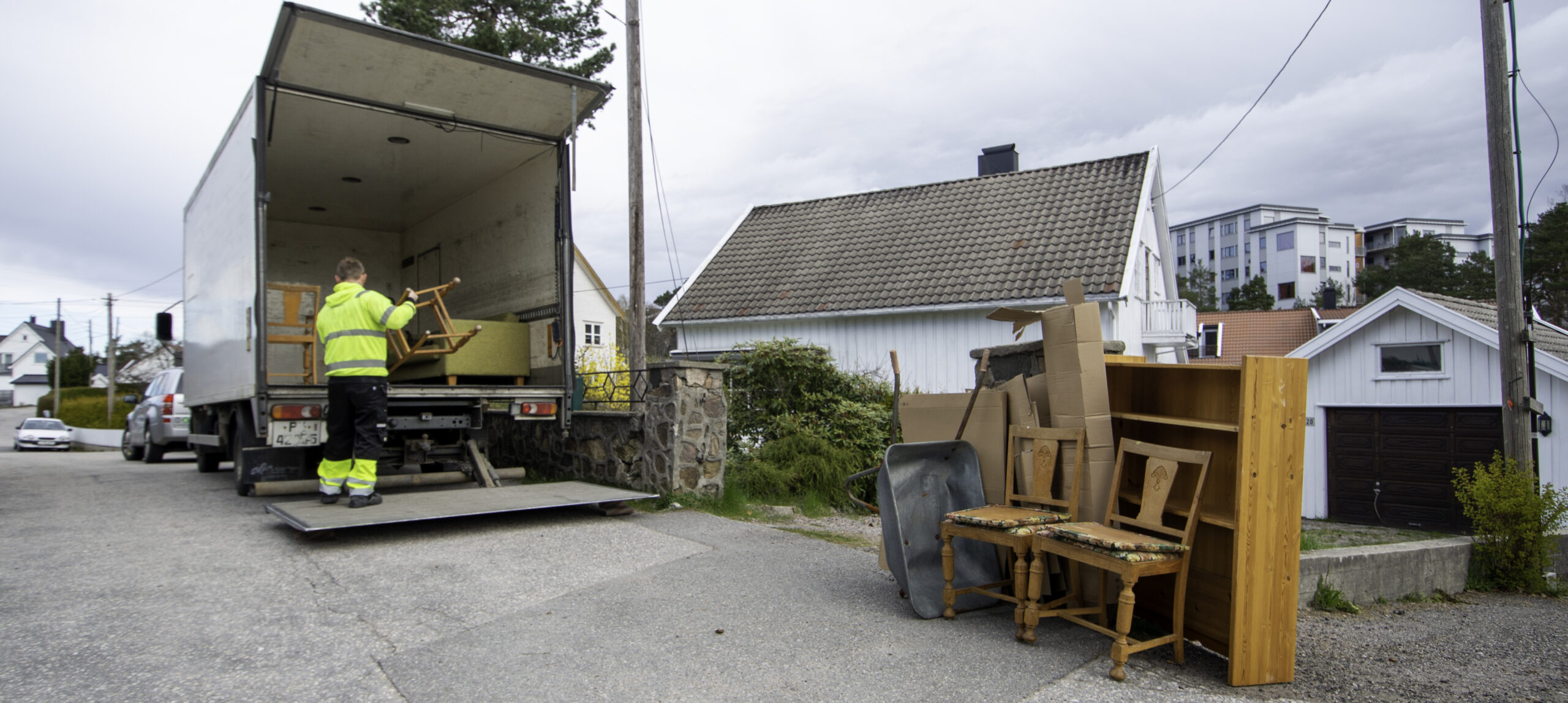 Mann i gult arbeidstøy som stabler inn møbler i en varebil, mens det står igjen møbler, trillebår og pappemballasje på gaten utenfor ut hus