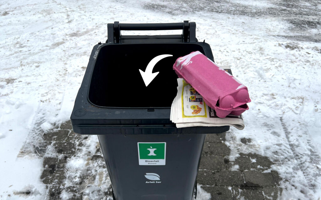 Åpen bioavfallsdunk om vinteren. Avis og eggekartong plassert på kanten, med pil som peker nedi dunken.